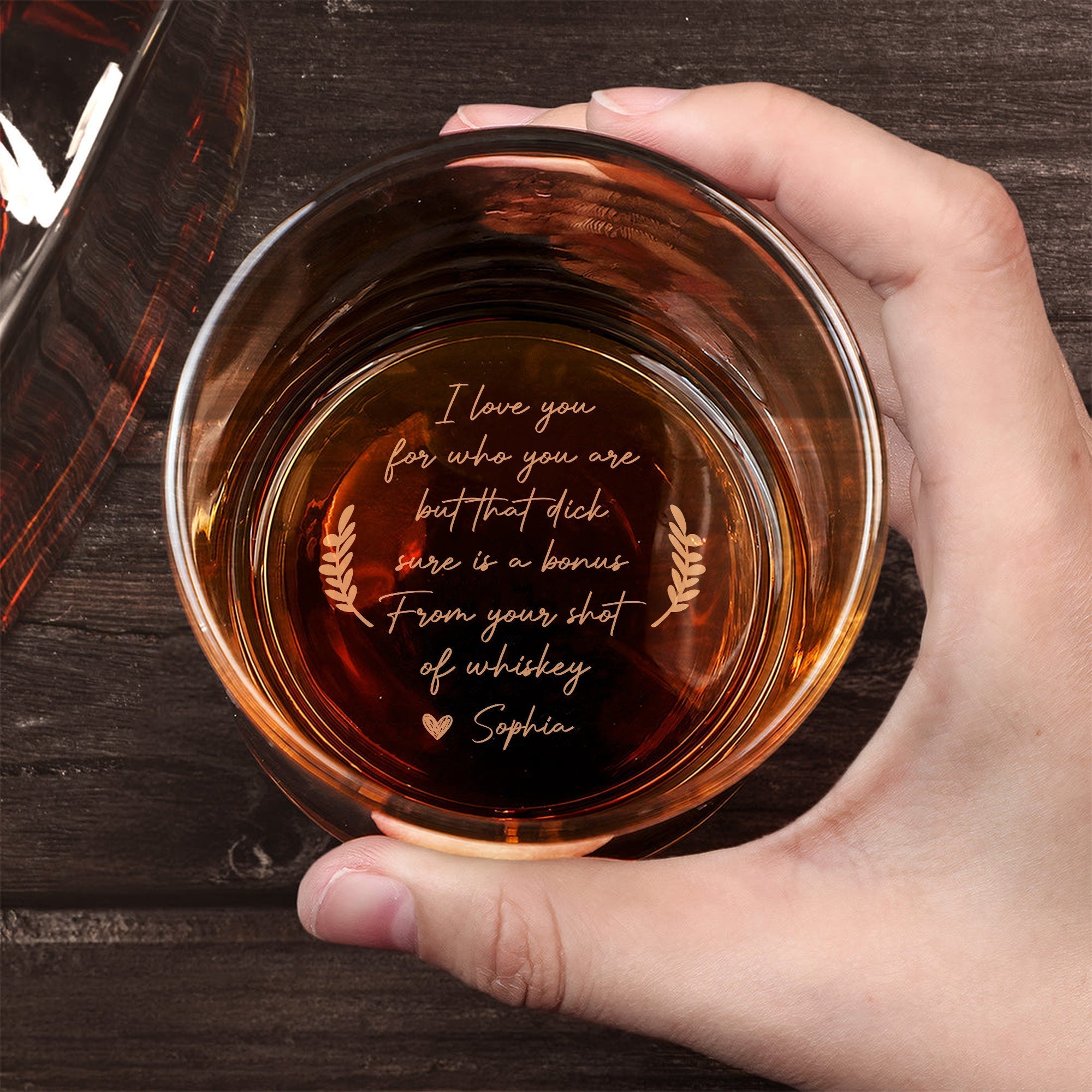 Personalized Whiskey Glass top view held in hand with amber whiskey and cheeky engraved love message at bottom on wooden table